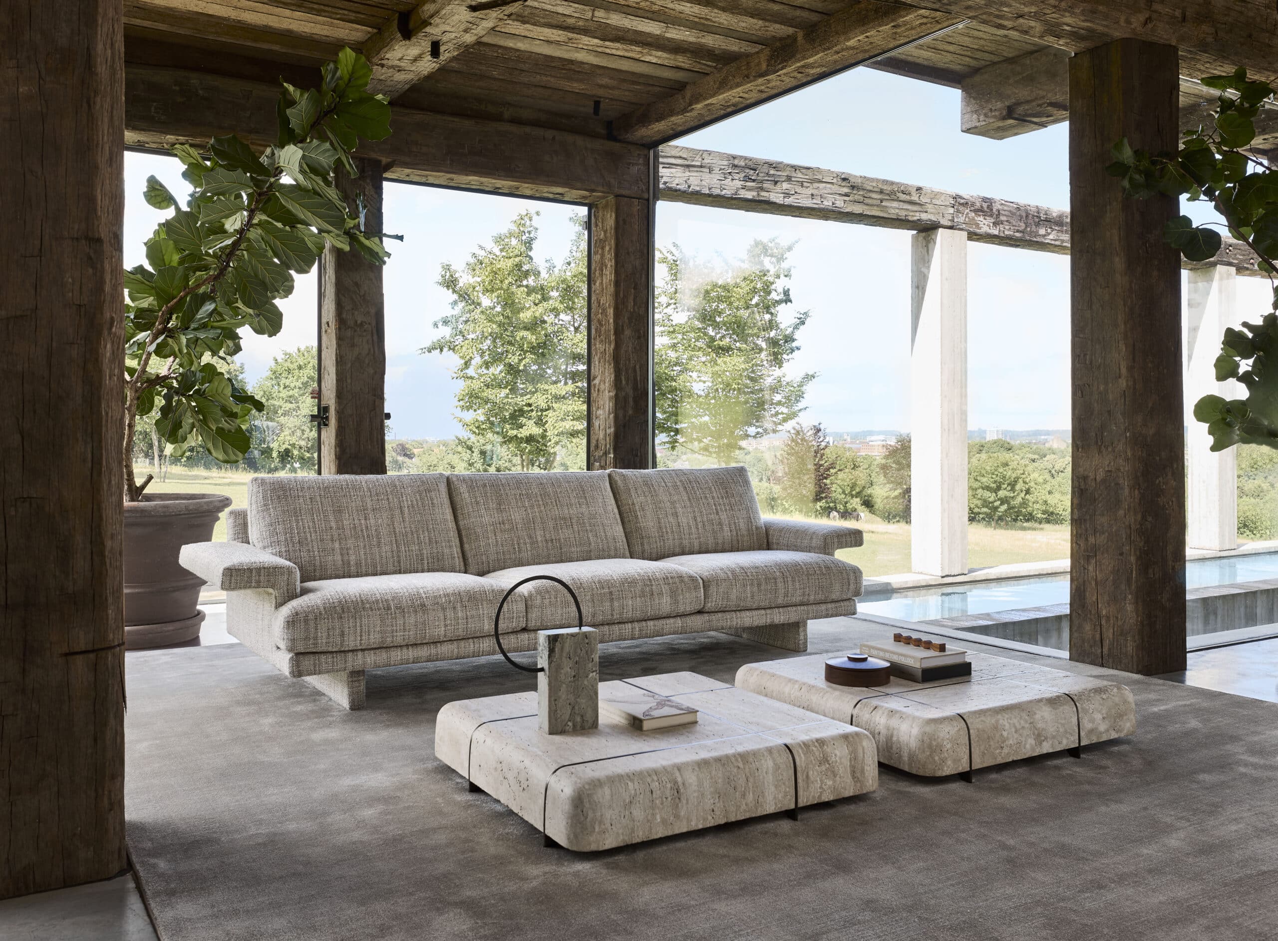 Linteloo fully opholstered Frank sofa and travertin stone Tessera coffee tables. The fabric of the sofa is textured. Shown in a living room with a lot of glass and robust wooden beams.